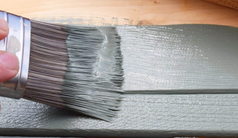 A close up of the bristles of a wide decorator’s paint brush coated with green paint as it’s used to paint a wood surface