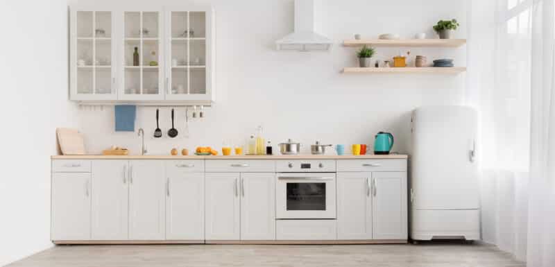 Interior of modern comfortable kitchen. Multicolored cups and teapot, orange juice in glass and utensils on white furniture, refrigerator, flowers in pots on shelves, light wall in daylight, panorama